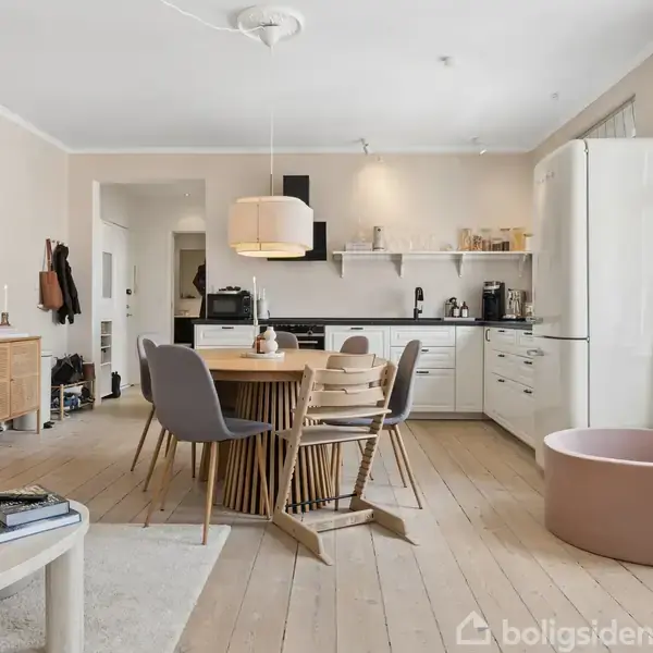 Spisebord med stole står i lyse, moderne åbent køkken/stue. Rummet har trægulv, hvidt køkken, køleskab og vindue mod højre. En beige sofa og sofabord ses til venstre.