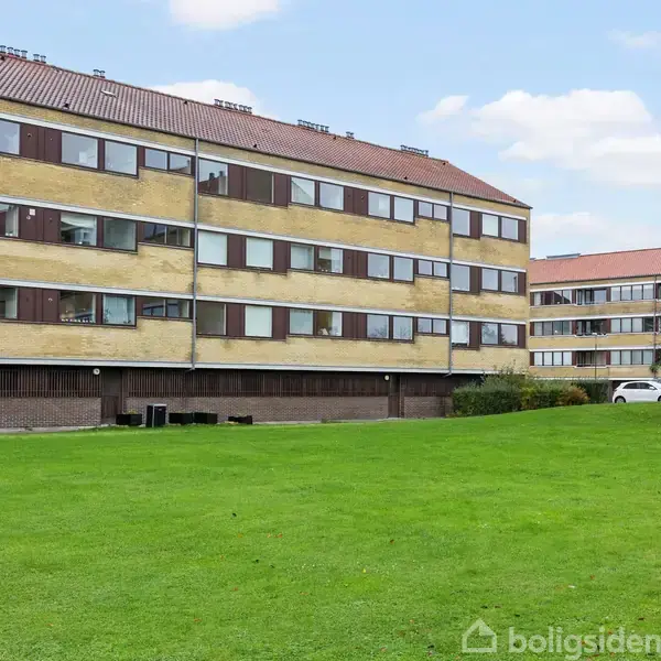 Flere gule murstensbygninger med røde tage står i en række. Foran bygningerne spreder en grøn græsplæne sig i et åbent område under en blå himmel med få skyer.