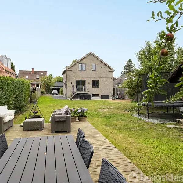 Et hus ses i baggrunden med en terrasse. I forgrunden findes en træterrasse med sorte havemøbler, omgivet af en grøn have med græsplæne, hegn og træer.