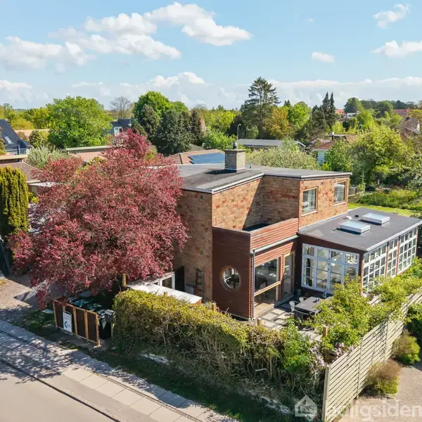 Et moderne hus med murstensfacade og store vinduer er omgivet af frodig have og langs en stille villavej. Et stort træ med røde blade står ved siden af huset.