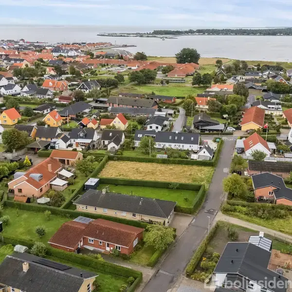Flere huse står rolige i en velordnet bydel, omgivet af grønne områder nær vandet. En krøllet vej snor sig gennem kvarteret, mens havet skaber en rolig baggrund.