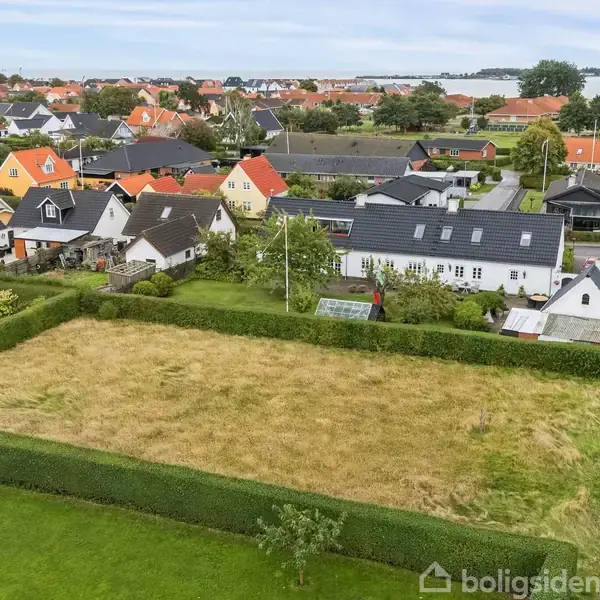 En tom grund ligger stille i en parcelhuskvarter, omkranset af hække. I baggrunden ses farverige huse og et glimt af havet.