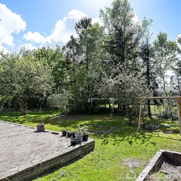 En have med en terrasse af stenfliser med potteplanter, en gynge hængende fra en træramme og omkringliggende træer under en klar blå himmel med få skyer.