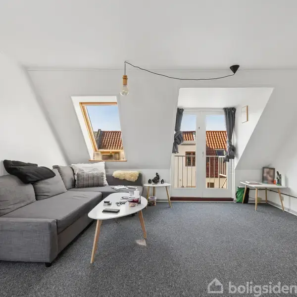 Grå sofa står i stue med skråvægge, garder bøger og pyntepuder, ved siden af bord med blade og fjernsyn. Store vinduer viser tagene udenfor, lysindfald oplyser rummet.