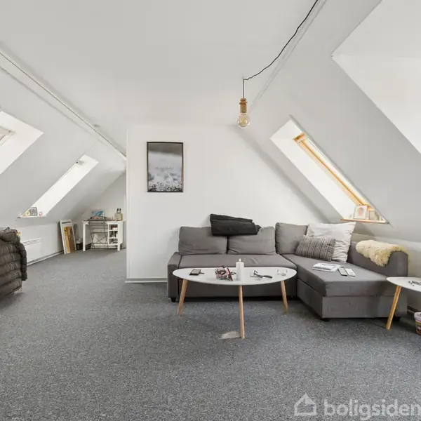 En grå sofa med hvide sofaborde står i en lys stue med skråvægge og loftvinduer. Et spisebord ses i baggrunden.