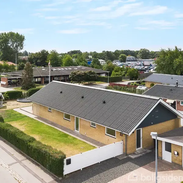 En enkeltstående, gul murstensvilla med sort tag ses fra luften. Huset ligger i et boligområde med velplejede grønne hække og flere nabohuse omgiver det i baggrunden.