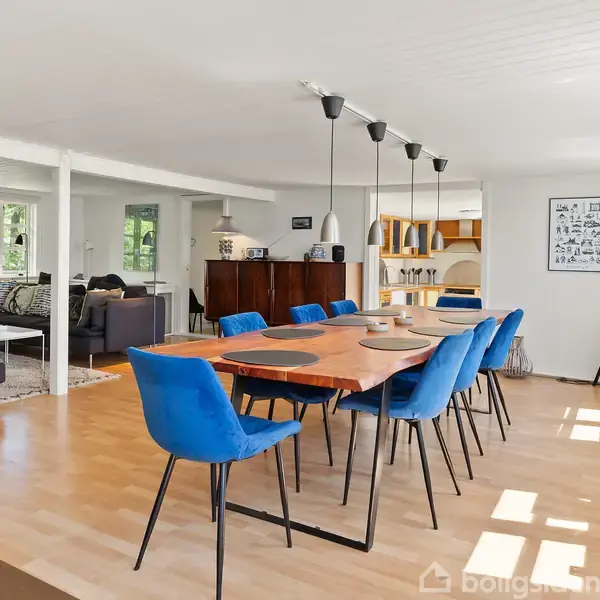 Spisebord med blå stole står i et lyst, åbent rum med parketgulv. I baggrunden ses en sofagruppe, skænk og store vinduer mod en have. Lamper hænger over bordet.