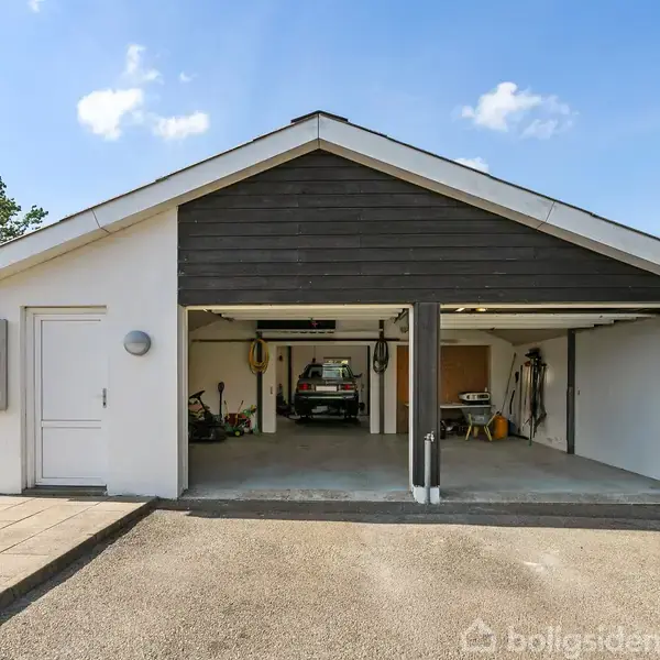 En hvid garage med to åbne porte. Indeni er der en bil og diverse værktøj opbevaret. Beliggende i en have med græsplæne under en blå himmel med skyer.