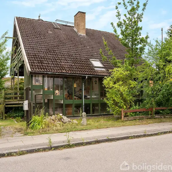 Et halvt dobbelthus med brun tegltag og en stor glaskarnap. Det står i en grøn have med små træer og buske. Der er en lille sti og en postkasse ved huset.