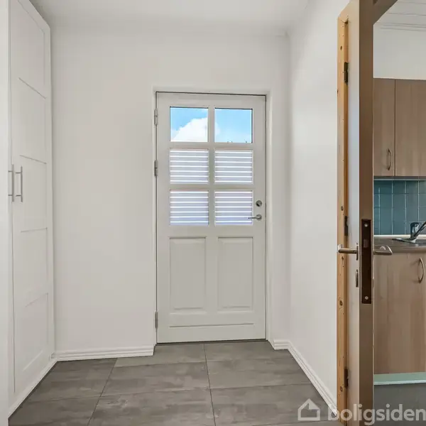 Dør står åben mod køkkenet, omgivet af hvide vægge. En hvid yderdør med vinduer er i baggrunden. Fliser dækker gulvet, skabe til venstre.