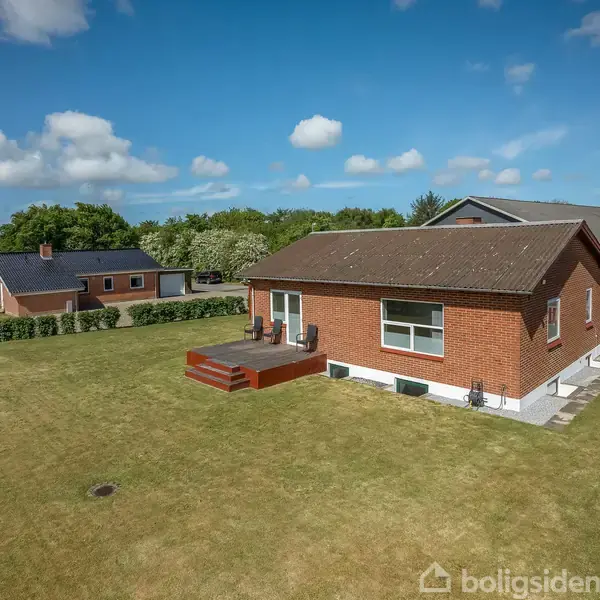 En rød murstenshus med en terrasse står på en stor græsplæne. Bag huset ses flere træer og en blå himmel med hvide skyer.