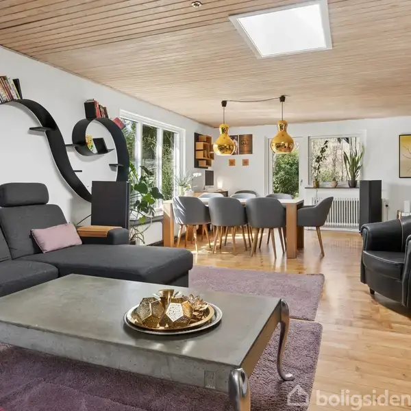 Hvidt hus med carport, delvist skjult af træer, står bag en hvid stakit. Græsset og en jordvej leder til indgangen. Træer omgiver huset på en overskyet dag.