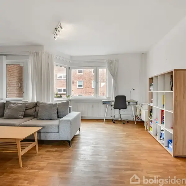 En grå hjørnesofa står i en lys stue med trægulv. Der er et sofabord foran sofaen, en reol til højre, og et skrivebord ved vinduet.