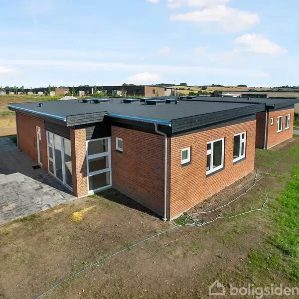 Et moderne, fladt tagstenhus står stille på en nyudviklet grund med græs. Huset er bygget af rød mursten og har flere vinduer. Området omkring huset er åbent og landligt.