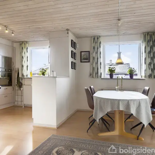 Et rundt spisebord dækket med hvid dug står ved to vinduer med grå gardiner. En lampe hænger over bordet. Til venstre er et åbent køkken med hvide skabe.
