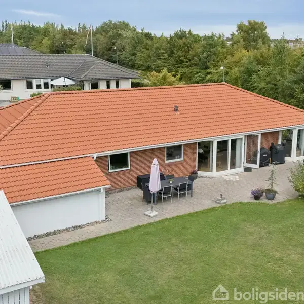 En rødt teglhustag dækker et enkelt etplanshus med store vinduer og en terrasse. Haven er velholdt og omfatter et drivhus og diverse planter, omkranset af grønne træer.