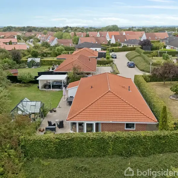 Et hus med rødt tag står omgivet af en velplejet have med grønt hegn. Til venstre ses et drivhus og terrasse; i baggrunden strækker et boligkvarter sig med lignende huse.