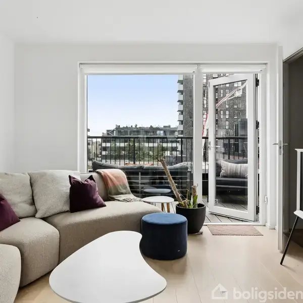 En lys stue har en beige sofa med puder, et hvidt sofabord, en fladskærms-tv på stativ, og en åbnet alubalkondør, som fører til en altan med udsigt over moderne bygninger.
