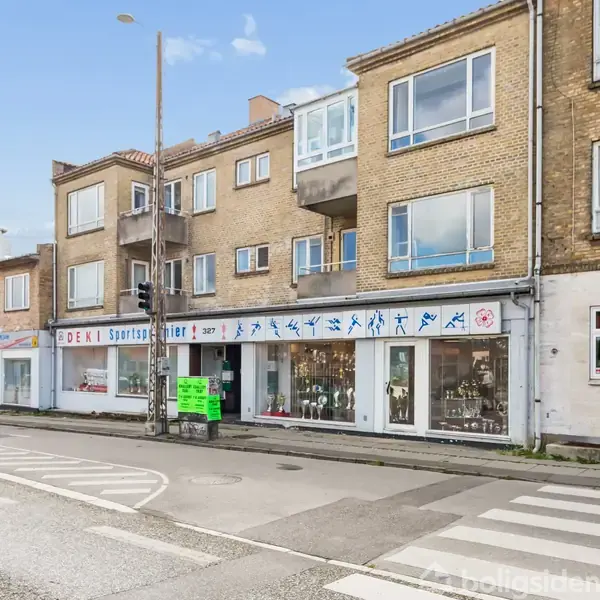 En gul murstensbygning med to etager, der huser "DEKI Sportstøj" butik. Store vinduer viser sportsudstyr. Til venstre en vej med fodgængerfelt under blå himmel.