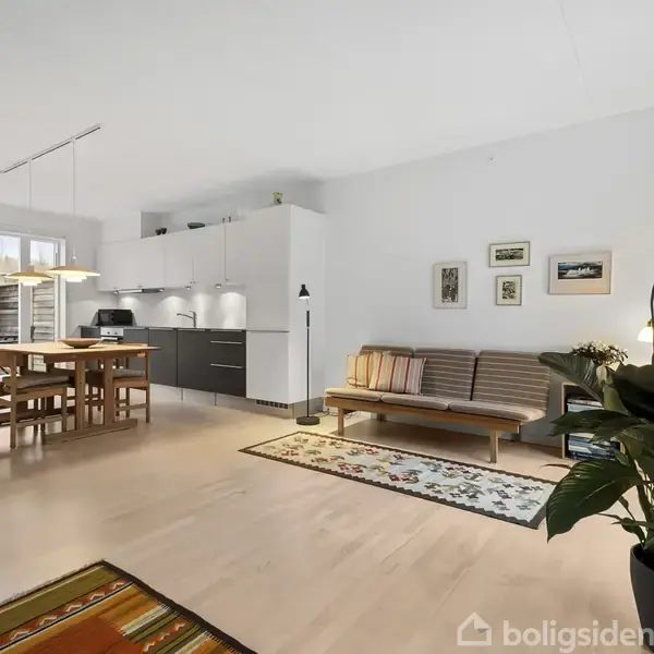 Stue med trægulv og sofa til højre, spisebord med stole placeret til venstre nær køkken. Indendørs planter og vægkunst tilføjer dekoration.