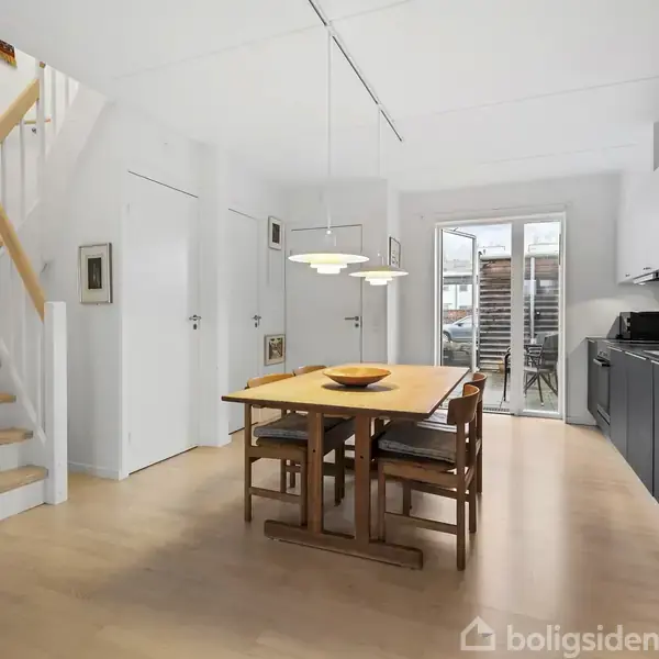 Træbord med fire stole står i moderne køkken med hvide vægge og skabslåger. Rummet har trægulv, trappe til venstre og glasdør mod terrasse. Loftslampe hænger over bordet.