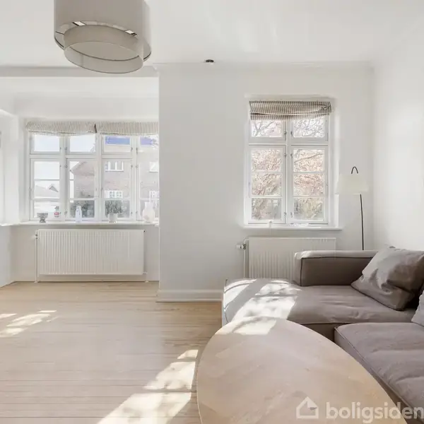 En brun sofa står i et lyst rum med trægulv. Sollys strømmer ind gennem store vinduer, og en lampe hænger i loftet. En reol med legetøj ses til venstre.