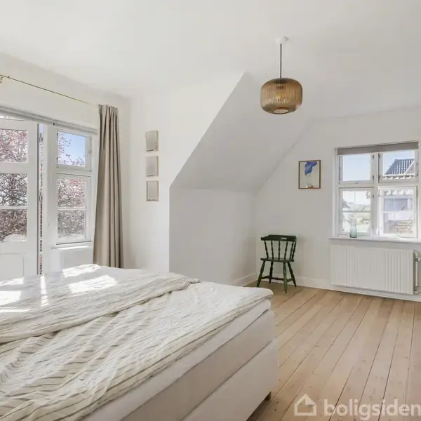 Soveværelse med seng til venstre, sollys strømmende ind gennem glasdør. Hvidmalede vægge og trægulv. Grønt træstol ved vindue. Brunt loftslampe og klædeskab til højre.
