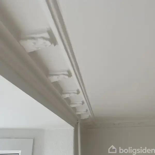 En hvid loftsliste med dekorative elementer er monteret langs en væg. Listens detaljer giver et elegant udseende i det minimalistiske rum.