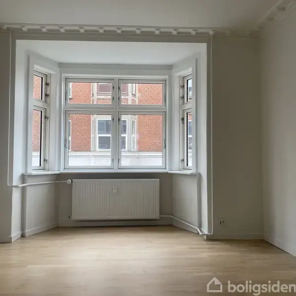 Et lyst værelse med et stort karnapvindue; vinduet består af flere mindre ruder. Værelset har lyse trægulve, og en radiator er placeret under vinduet. Udsigt til nabobygning.