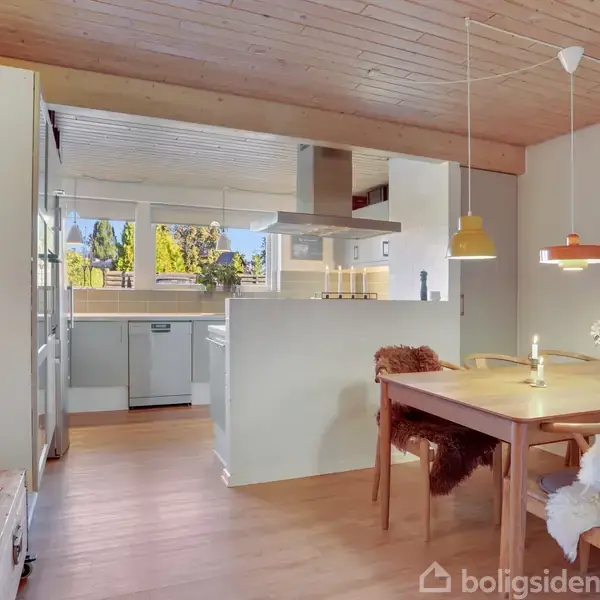 Køkken med åbent spiseområde. Træbord og stole pyntet med skind under flere farvede lamper. Vidt udsyn gennem store vinduer. Lyse trægulve og hvide vægge omkranser rummet.