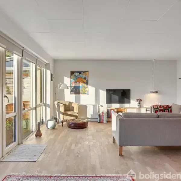 Stue med sofa og lænestol, hvor solen skinner ind gennem store vinduer. Kunstværker hænger på væggene. En fladskærms-TV er placeret ved siden af sofaen.