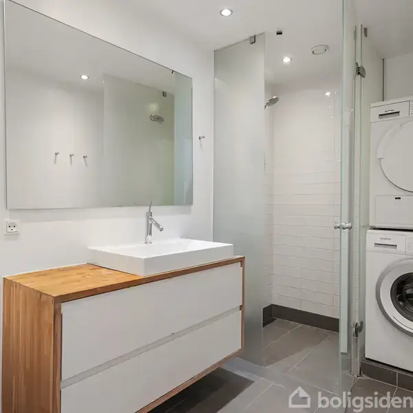 Lyst lejlighedskompleks med træer foran, set fra en kanal med fortøjede både. Billedet viser en moderne urban boligstil med gade og reflekterende glasfacader i baggrunden.