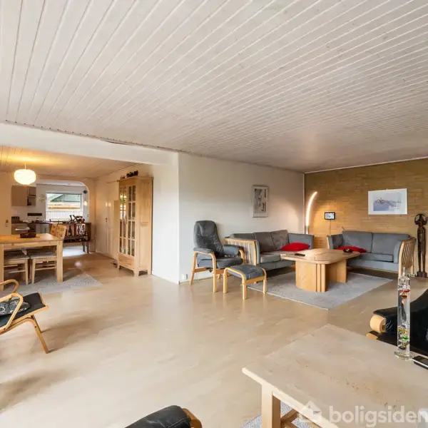 Stue med lædersofaer og stole omkring et sofabord; i baggrunden en spisestue med spisebord og stole. Rummelig, lys indretning med træloft og parketgulv.