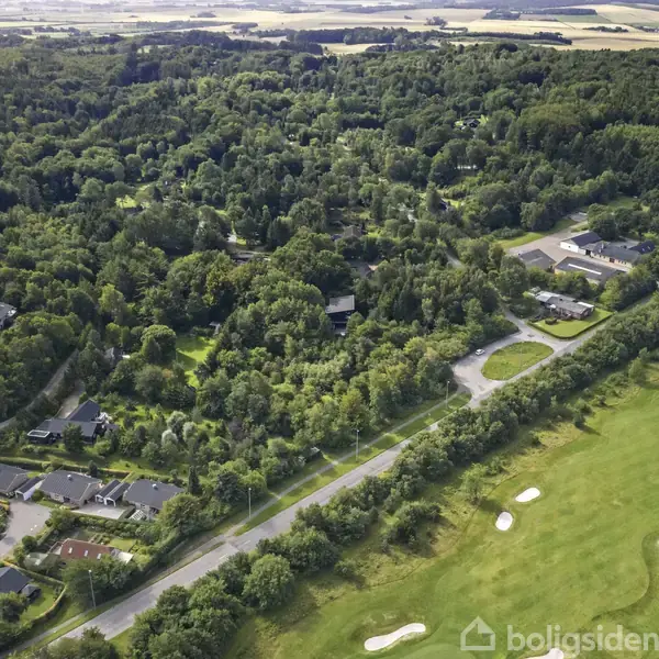 En skov fyldt med tæt bevoksning omgiver et boligområde, hvor huse ligger langs en vej. Til højre ses en velplejet golfbane med sandbunkers og åbne græsarealer.