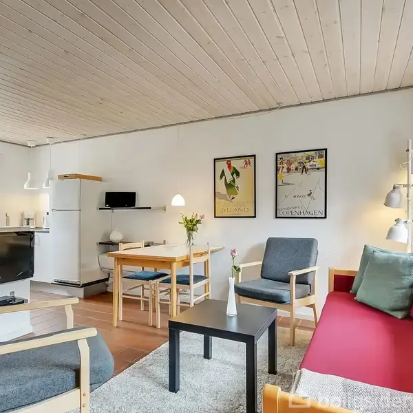 En stue med sofa og stole arrangeret omkring et sofabord. Et spisebord med stole står ved siden af køkkenområdet, hvor der er et køleskab og TV. Vægplakater hænger ved siden af hinanden.