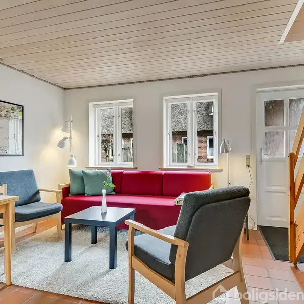 En stue med en rød sofa, der står langs en væg med tre vinduer. To stole og et sofabord står foran sofaen. En trappe til højre fører opad.