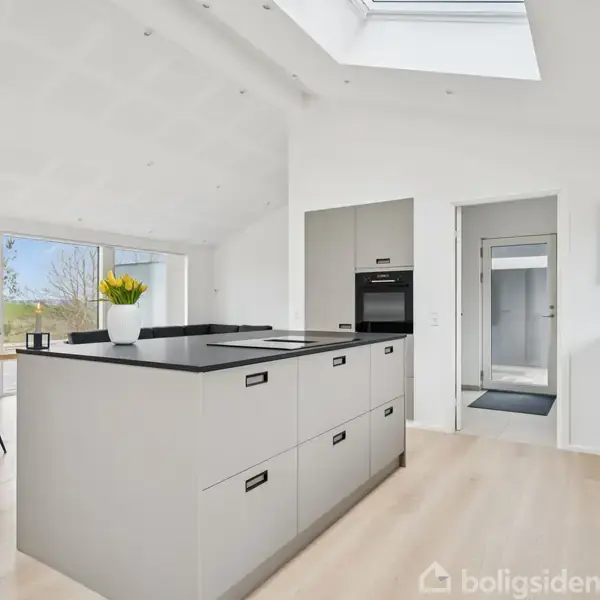 Et moderne køkken med en stor grå køkkenø, en integreret ovn og lyse trægulve. I baggrunden er der et spisebord ved panoramavinduer, der viser en grøn udsigt.