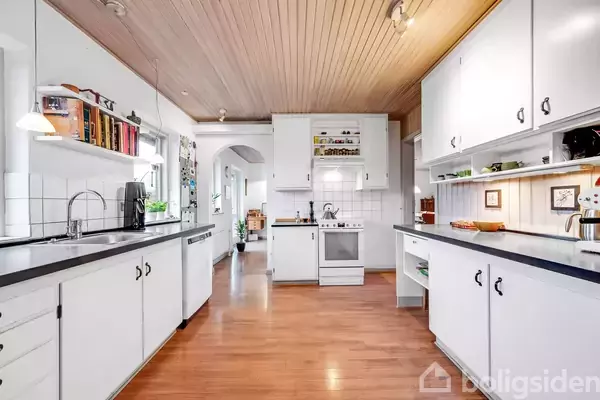 Køkkenet har hvide skabe og sorte bordplader, trælaminatgulv, og hvidt fliset backsplash. En vask og opvaskemaskine er til venstre, mens en komfur er i midten. Bøger og dekorationer pryder hylderne.