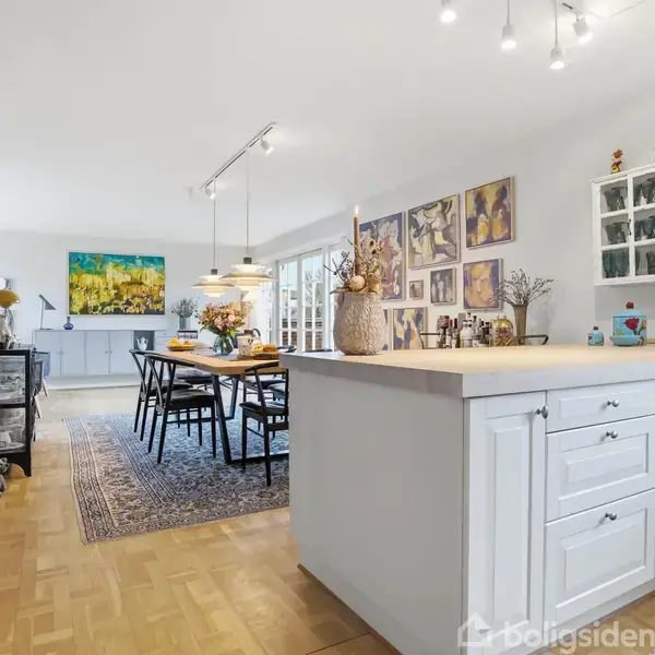 Et køkken med en stor, hvid køkkenø, omgivet af malerier på væggen. Et spisebord med stole står over et tæppe, og der er lamper hængende fra loftet.