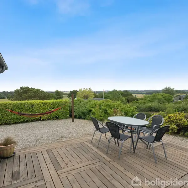 En terrasse med et bord og fire stole står på træplanker. En hængekøje hænger i baggrunden, omgivet af buske og natur. Himmel er klar og blå.