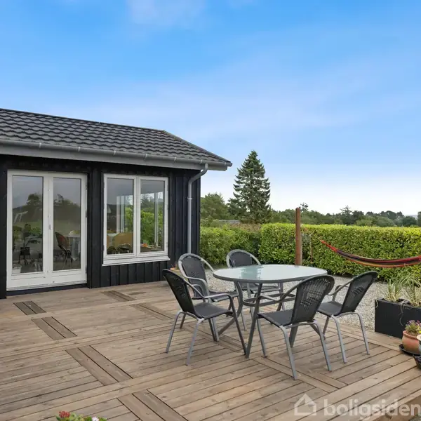 Et udendørs terrasseområde med et rundt glasbord og seks stole står på trædæk. Til venstre ses en grill, og i baggrunden er der grønne hække og et skovområde.