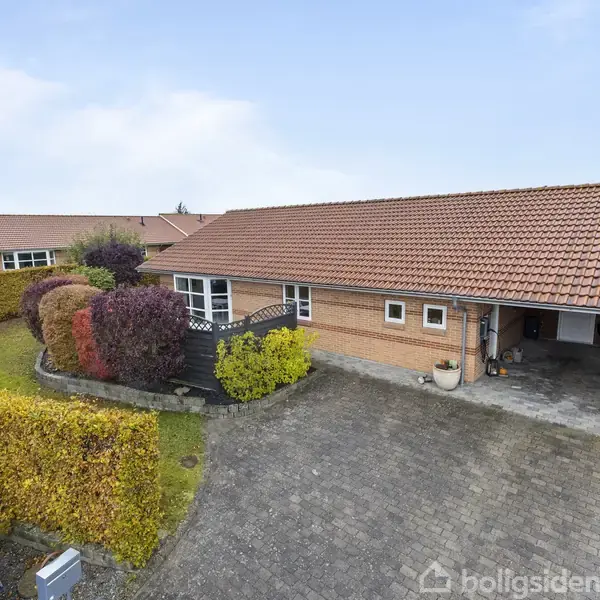 Et murstenshus med rødt tegltag står i en velplejet have med farverige buske. Foran er en gårdsplads med fliser og en carport. Himmel er let overskyet.