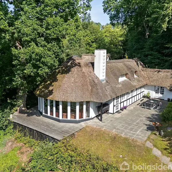 Et stråtækt hus ligger i en skovklædt have med terrasser i sten. Huset har et rundt vinduesparti, og skorstenen bærer teksten "Lyngager." Træer omgiver den stille ejendom.