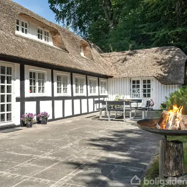 Bindingsværkshus med stråtag står stille ved siden af et flammebål i en bålplads. Det ligger på en flisebelagt terrasse omgivet af frodig skov.