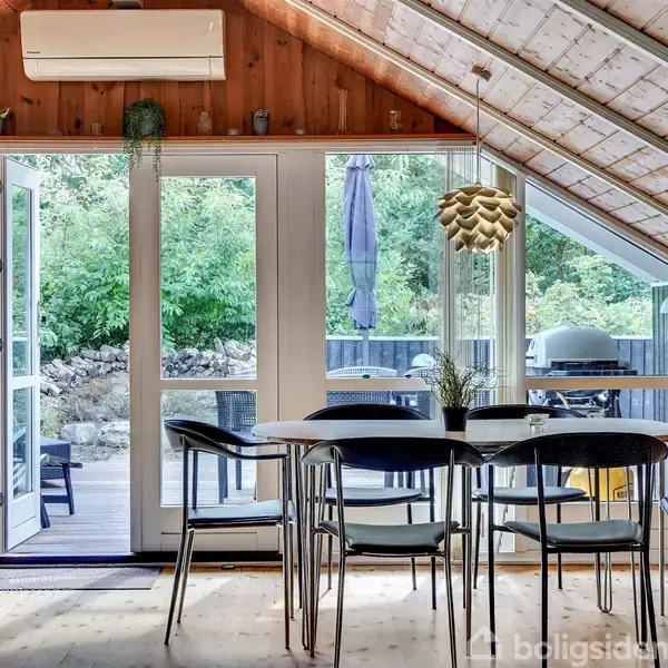 Et spisebord med seks stole står foran glasdøre, der åbner ud til en terrasse. Omkring er træpaneler og et skråt loft. Udenfor ses en grill og grønne buske.