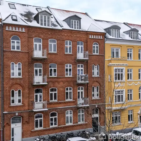 Række af klassiske bylejligheder med røde og gule facader, små altaner og sneklædte tage, omgivet af cykler og træer i en bymæssig gade.