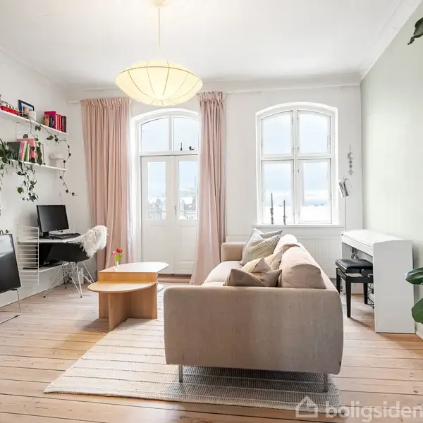 Sofa står i en stue med lyse trægulve. Et bord, en stol ved arbejdsstation og en fladskærm er placeret foran vinduer med lyserøde gardiner. Grønne planter pynter rummet.