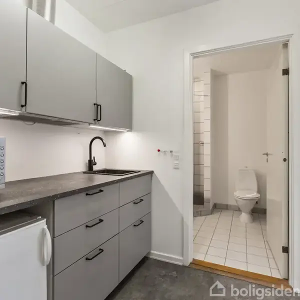 Et moderne køkken med grå skabe og skuffer, en mikroovn på bordpladen og en sort vandhane. I baggrunden ses en åben dør til et badeværelse med et toilet.