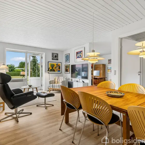 En lys stue med spisebord og seks stole i forgrunden. En sort lænestol med skammel står ved en skænk til venstre. Vægge dekoreret med malerier; dør ud til terrasse.