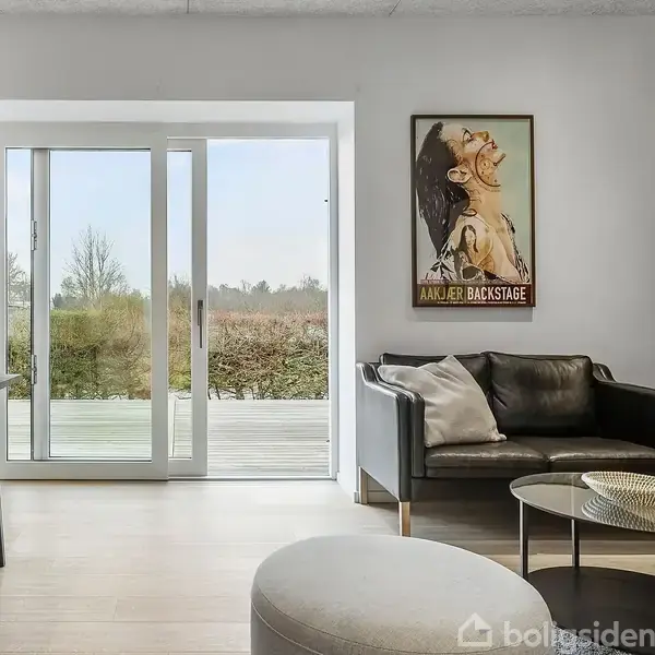 Stue med sort sofa og rund sofabord, som står foran en stor glasdør med udsigt til en have. To kunstplakater hænger på væggene med teksten "JACKIE LOVE" og "ANKER BACKSTAGE."